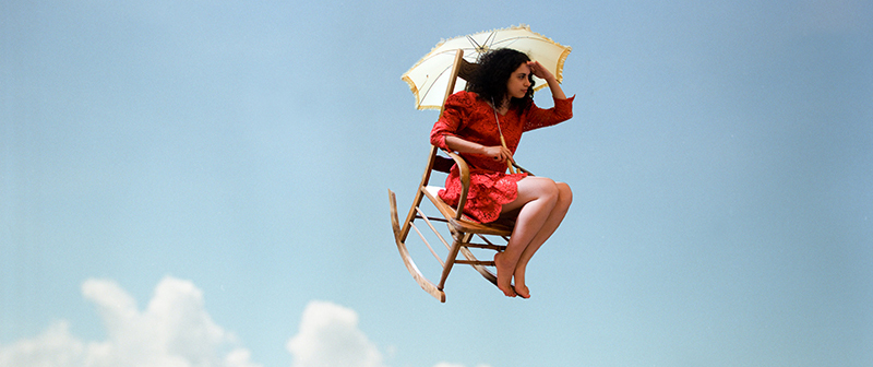 a photo of a girl in a red dress and yellow parasol flying purposefully through the air in a rocking chair