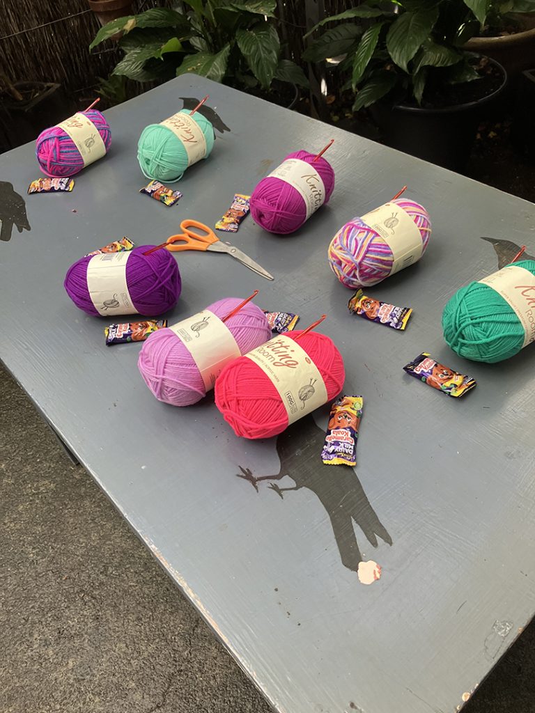 a table with painted crows and balls of coloured yarn, each with a crochet hook. wrapped Caramello Koalas dot the table.