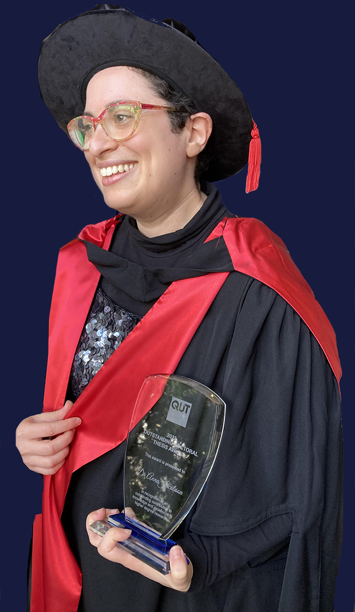 Anna smiling in her graduation gown and holding the outstanding doctoral thesis award, a glass trophy
